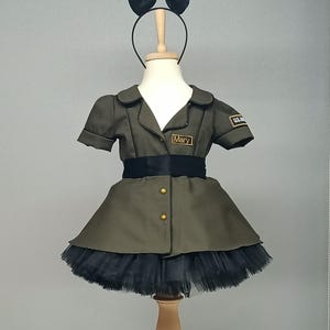 May include: Olive green military-style dress with a black tulle skirt, black bow headband, and a black belt. The dress has gold buttons and a name tag that reads "Mary". A "U.S. Army" patch is visible on the sleeve.