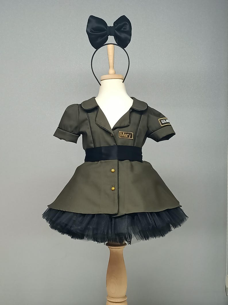 May include: Olive green military-style dress with a black tulle skirt, black bow headband, and a black belt. The dress has gold buttons and a name tag that reads "Mary". A "U.S. Army" patch is visible on the sleeve.