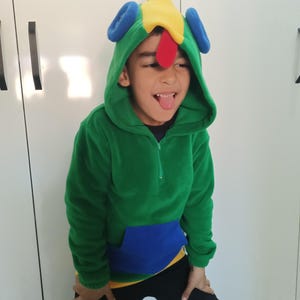 Puede incluir: Un ni&ntilde;o lleva una sudadera con capucha verde, azul y amarilla con una cabeza de gallo colorida en la capucha. El ni&ntilde;o est&aacute; sacando la lengua.