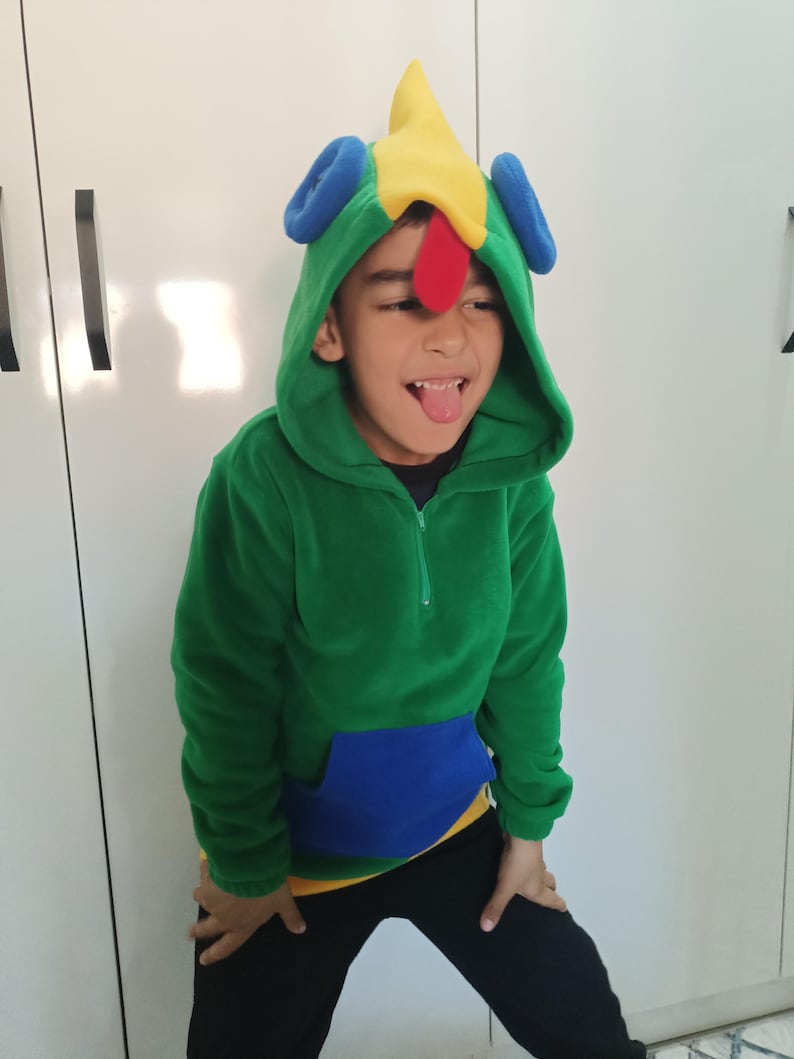Puede incluir: Un ni&ntilde;o lleva una sudadera con capucha verde, azul y amarilla con una cabeza de gallo colorida en la capucha. El ni&ntilde;o est&aacute; sacando la lengua.