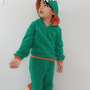 Peut inclure: Un enfant portant un costume de dinosaure vert et orange avec une queue. Le costume comprend un sweat &agrave; capuche et un pantalon assorti.