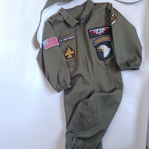 May include: Olive green Top Gun Maverick flight suit with patches, including U.S. Air Force and Airborne. Includes a matching aviator hat with goggles. The jumpsuit has a zipper closure and elastic cuffs at the wrists and ankles.