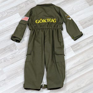 May include: Olive green jumpsuit with multiple pockets, a collared neckline, and elastic waist. The back features the word "G&Ouml;KTUĞ" in yellow lettering, along with an American flag patch and Army insignia. The jumpsuit has long sleeves and a button closure.