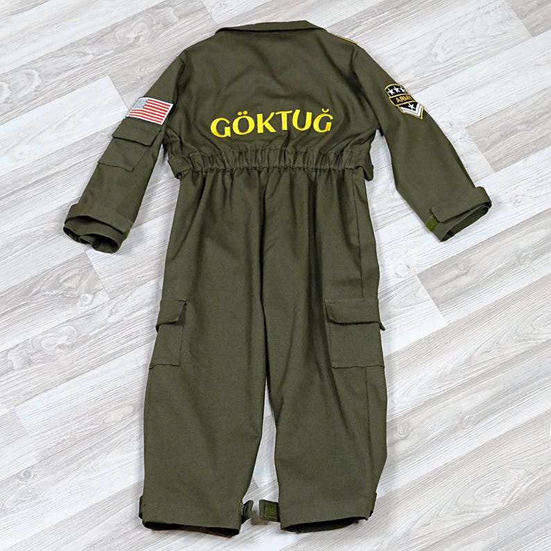 May include: Olive green jumpsuit with multiple pockets, a collared neckline, and elastic waist. The back features the word "G&Ouml;KTUĞ" in yellow lettering, along with an American flag patch and Army insignia. The jumpsuit has long sleeves and a button closure.