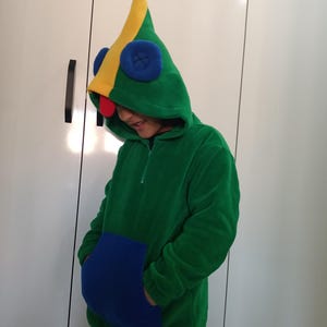 Puede incluir: Una sudadera con capucha verde con una capucha puntiaguda amarilla y verde. La capucha tiene un bot&oacute;n azul con una X y una nariz roja. La sudadera tiene un bolsillo azul y una banda amarilla en la parte inferior.