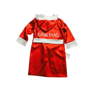 May include: A red satin boxing robe with white trim on the hood, cuffs, and belt. The name "G&Ouml;KTUĞ" is embroidered in white on the back. The robe is designed for a child.