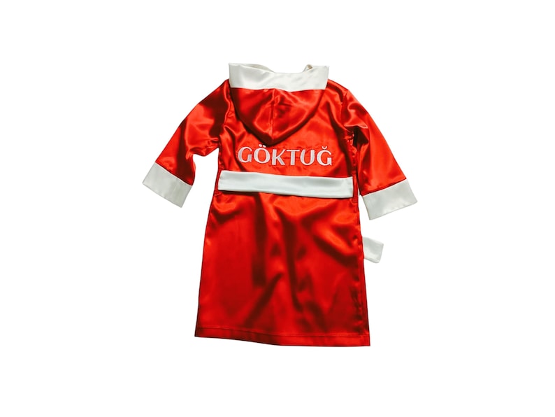 May include: A red satin boxing robe with white trim on the hood, cuffs, and belt. The name "G&Ouml;KTUĞ" is embroidered in white on the back. The robe is designed for a child.