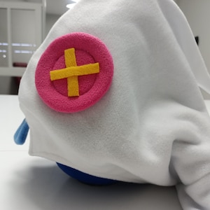 May include: A white plush hat with a pink circle and yellow cross emblem. The hat has a blue accent and a rounded shape. The hat is made of soft, fuzzy material.