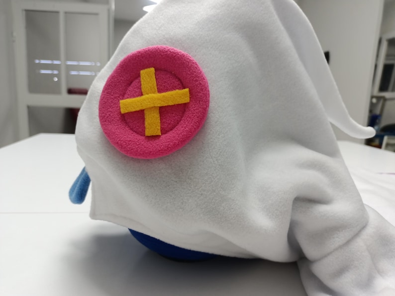 May include: A white plush hat with a pink circle and yellow cross emblem. The hat has a blue accent and a rounded shape. The hat is made of soft, fuzzy material.