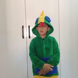 Puede incluir: Un ni&ntilde;o lleva una sudadera con capucha verde, amarilla y azul con una capucha en forma de p&aacute;jaro. La capucha tiene un pico rojo y ojos azules.