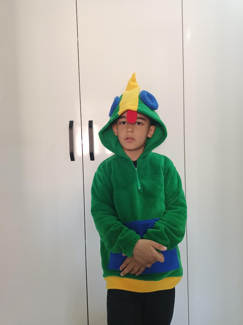 Puede incluir: Un ni&ntilde;o lleva una sudadera con capucha verde, amarilla y azul con una capucha en forma de p&aacute;jaro. La capucha tiene un pico rojo y ojos azules.