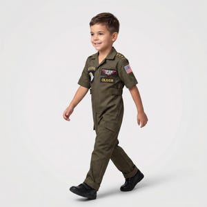 May include: Olive green Top Gun flight suit with patches and the name "OLIVER". The costume includes a collared short-sleeve jumpsuit with pockets and a US flag patch. The child is wearing black shoes.