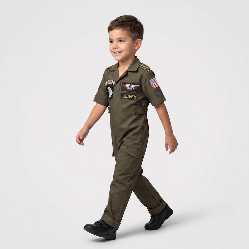 May include: Olive green Top Gun flight suit with patches and the name "OLIVER". The costume includes a collared short-sleeve jumpsuit with pockets and a US flag patch. The child is wearing black shoes.
