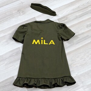 Puede incluir: Un vestido verde con un dobladillo con volantes y mangas cortas. El vestido tiene el nombre "MILA" bordado en la espalda con hilo amarillo.