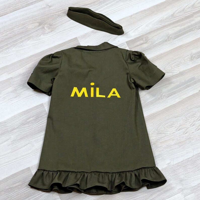 Puede incluir: Un vestido verde con un dobladillo con volantes y mangas cortas. El vestido tiene el nombre "MILA" bordado en la espalda con hilo amarillo.