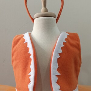 May include: Orange and white fox costume for a child, featuring a tutu skirt, a vest with a white trim, and a headband with fox ears and a bow.