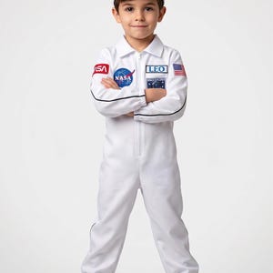 Może przedstawiać: Biały kostium astronauty dla dzieci, z naszywką z logo NASA, plakietką z imieniem "LEO" i naszywką z flagą amerykańską. Str&oacute;j zawiera pasującą czapkę i jest noszony z trampkami.