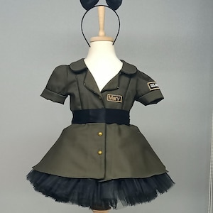 May include: Olive green dress with short sleeves, a black tulle skirt, and a black bow headband. The dress features gold buttons, a black belt, and a name tag that reads "Mary". A U.S. Army patch is visible on the sleeve.