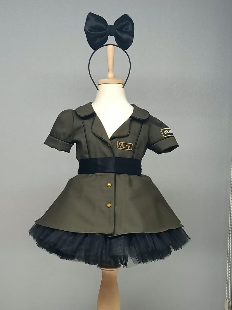 May include: Olive green dress with short sleeves, a black tulle skirt, and a black bow headband. The dress features gold buttons, a black belt, and a name tag that reads "Mary". A U.S. Army patch is visible on the sleeve.