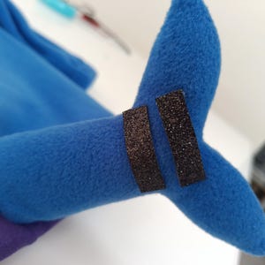 以下が含まれることがあります： 2つの黒いキラキラしたバンドが付いた、青いぬいぐるみのおもちゃの尻尾のクローズアップ。 尻尾は柔らかくふわふわした素材で作られています。 背景には、他の青い生地と白い表面がちらりと見えます。
