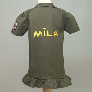 Puede incluir: Un vestido verde de manga corta con un dobladillo con volantes y un nombre bordado en oro "MILA" en la espalda. El vestido tiene un peque&ntilde;o parche de la bandera estadounidense en la manga izquierda.