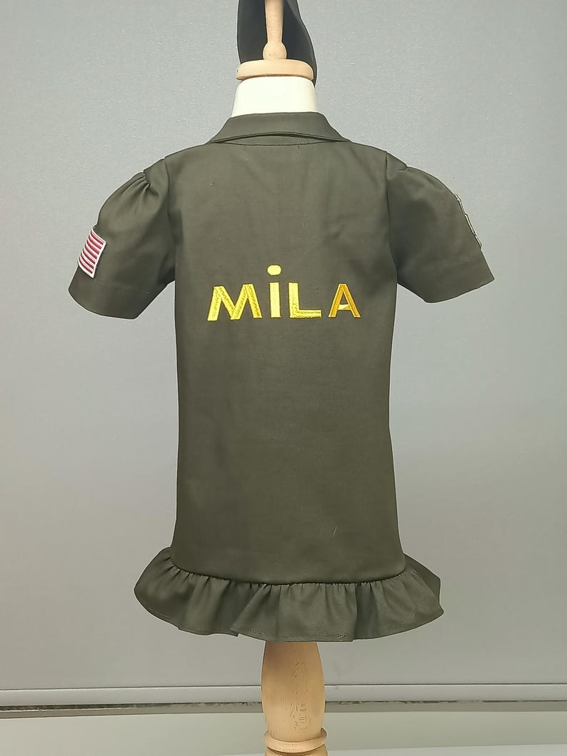 Puede incluir: Un vestido verde de manga corta con un dobladillo con volantes y un nombre bordado en oro "MILA" en la espalda. El vestido tiene un peque&ntilde;o parche de la bandera estadounidense en la manga izquierda.