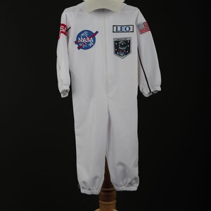 Może przedstawiać: Biały kostium astronauty NASA z pasującą czapką. Kombinezon posiada naszywki NASA i napis "LEO SPACE EXPLORER". Czapka ma logo NASA. Kostium jest wystawiony na drewnianym stojaku.