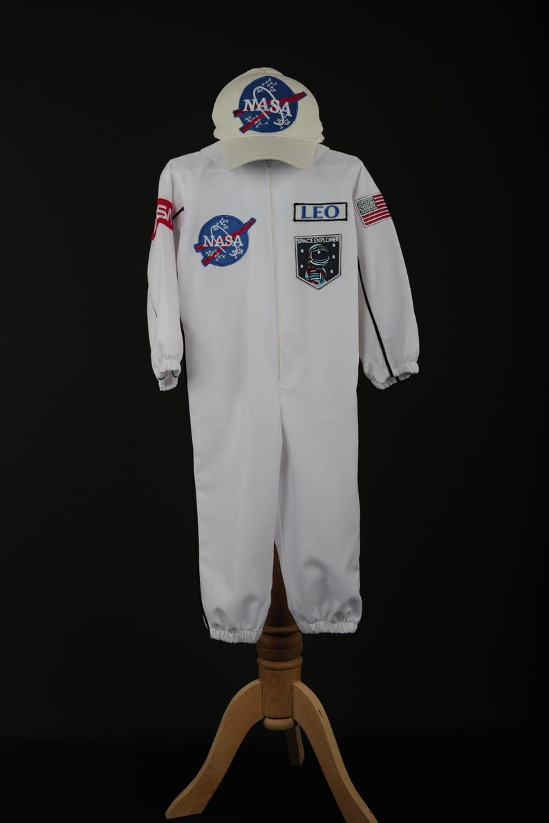 Może przedstawiać: Biały kostium astronauty NASA z pasującą czapką. Kombinezon posiada naszywki NASA i napis "LEO SPACE EXPLORER". Czapka ma logo NASA. Kostium jest wystawiony na drewnianym stojaku.