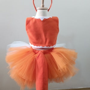 May include: Orange and white fox costume for a child. The costume includes a tutu skirt, a top with a white trim, and a tail. The costume is on a wooden stand.