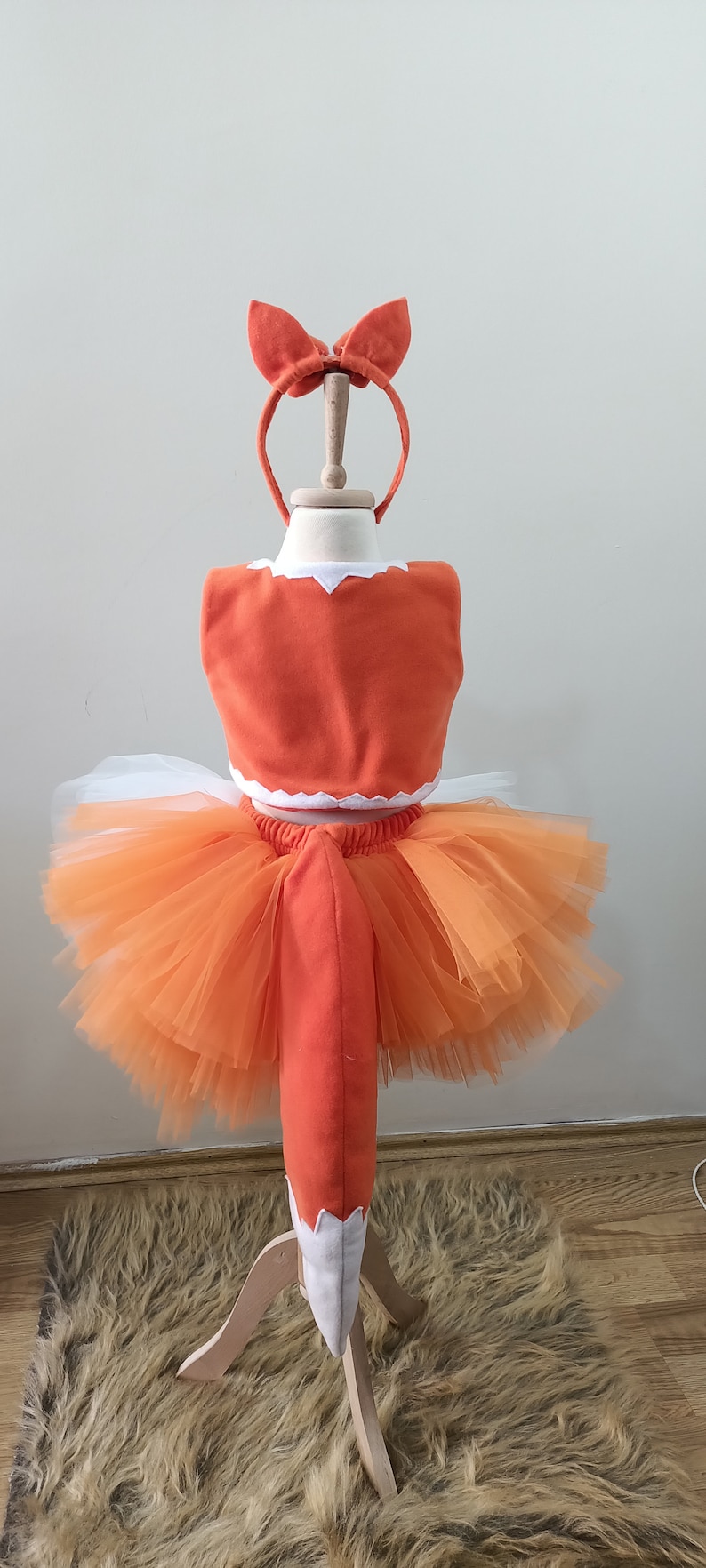 May include: Orange and white fox costume for a child. The costume includes a tutu skirt, a top with a white trim, and a tail. The costume is on a wooden stand.