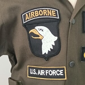 Puede incluir: Primer plano de una chaqueta de uniforme militar verde oliva. La chaqueta presenta parches que dicen "AIRBORNE" y "U.S. AIR FORCE". El parche "AIRBORNE" tiene un fondo negro con una cabeza de &aacute;guila blanca y un pico dorado. El uniforme tiene una botonadura.