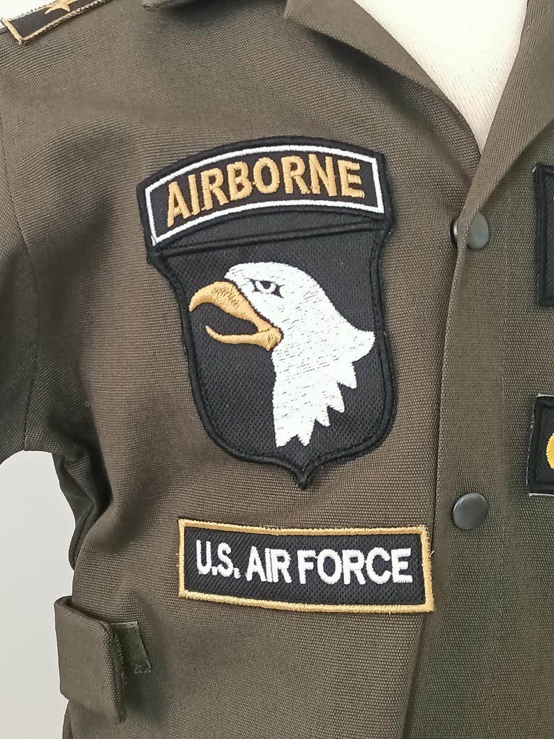 Puede incluir: Primer plano de una chaqueta de uniforme militar verde oliva. La chaqueta presenta parches que dicen "AIRBORNE" y "U.S. AIR FORCE". El parche "AIRBORNE" tiene un fondo negro con una cabeza de &aacute;guila blanca y un pico dorado. El uniforme tiene una botonadura.