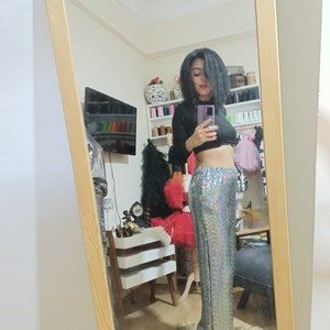 Puede incluir: Una mujer con un top negro de manga larga y pantalones plateados con lentejuelas. Est&aacute; de pie frente a un espejo y se est&aacute; tomando una selfie.