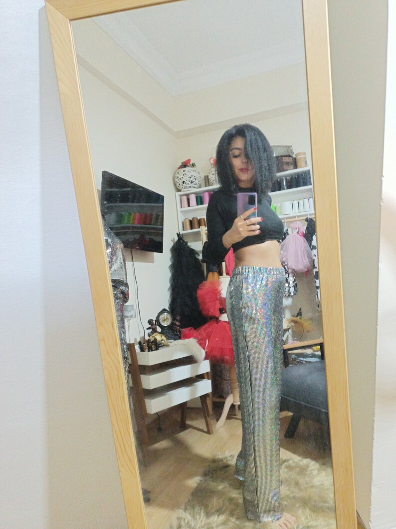 Puede incluir: Una mujer con un top negro de manga larga y pantalones plateados con lentejuelas. Est&aacute; de pie frente a un espejo y se est&aacute; tomando una selfie.