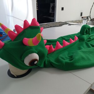 May include: A green dragon costume with pink and yellow accents. The costume features a detailed dragon head with large eyes, horns, and pink spikes along the back. The costume is made of soft fabric and is designed to be worn.