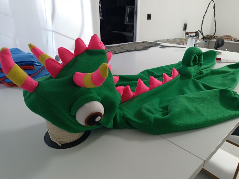 May include: A green dragon costume with pink and yellow accents. The costume features a detailed dragon head with large eyes, horns, and pink spikes along the back. The costume is made of soft fabric and is designed to be worn.