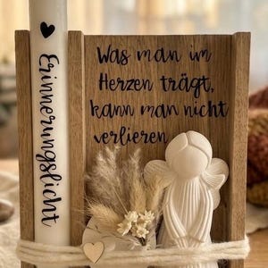 Könnte beinhalten: Ein Holzständer mit einer weißen Kerze, die ein schwarzes Herz und den Schriftzug "Erinnerungslicht" trägt, daneben eine weiße Engelsfigur und Trockenblumen. Das Holz ist mit dem Text "Was man im Herzen trägt, kann man nicht verlieren" graviert und mit einem dicken weißen Garn umwickelt.
