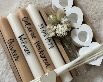 Puede incluir: Una bandeja de madera con cuatro velas cónicas en beige y blanco, con las inscripciones Günter, Wilma, Goldene Hochzeit y 31.05.1974. El conjunto incluye tres portavelas blancos en forma de corazón, un pequeño jarrón de cristal con flores secas y un envoltorio de hilo blanco con un pequeño corazón de madera.