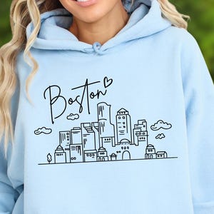 May include: Light blue hoodie with a black graphic of the Boston skyline and the word "Boston" in cursive. The hoodie has a front pocket and a drawstring hood. The design includes clouds and buildings.