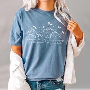 A person wearing a blue t-shirt with a mountain landscape design and a quote printed on it, along with a white jacket.