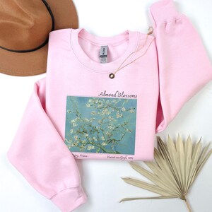 a pink sweatshirt with a graphic of a tree with white flowers, a brown hat, a necklace, a fan, and a plant on a white background.