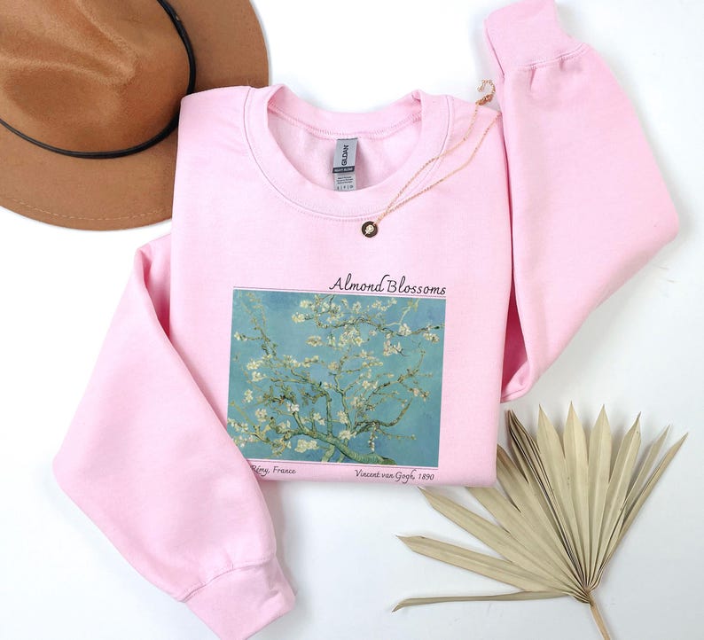 a pink sweatshirt with a graphic of a tree with white flowers, a brown hat, a necklace, a fan, and a plant on a white background.