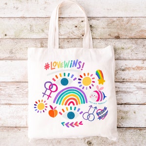 May include: A white tote bag with the text "#LOVEWINS!" and colorful illustrations including a rainbow, a sun, a bee, and an eye. The bag is on a white wooden background.