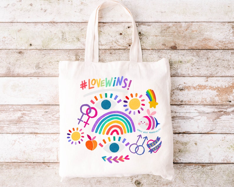 May include: A white tote bag with the text "#LOVEWINS!" and colorful illustrations including a rainbow, a sun, a bee, and an eye. The bag is on a white wooden background.
