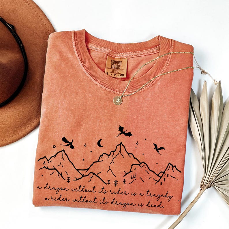 a pink t-shirt with a graphic design depicting mountains and a dragon flying over them, along with a quote that reads "A dragon without its rider is a tragedy. A rider without its dragon is dead." The t-shirt is laid out on a white surface, accompanied by a hat and a fan.