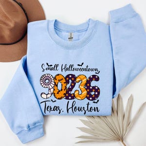 May include: A light blue sweatshirt with the text "Small Halloweentown 2023 Texas, Houston". The numbers are decorated with a spiderweb, bats, and orange and purple Halloween-themed patterns. A brown hat is visible in the background.