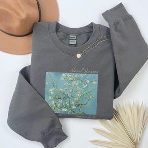 a gray sweatshirt with a graphic of a tree with white flowers, a brown hat, a necklace, and a fan on a white background.