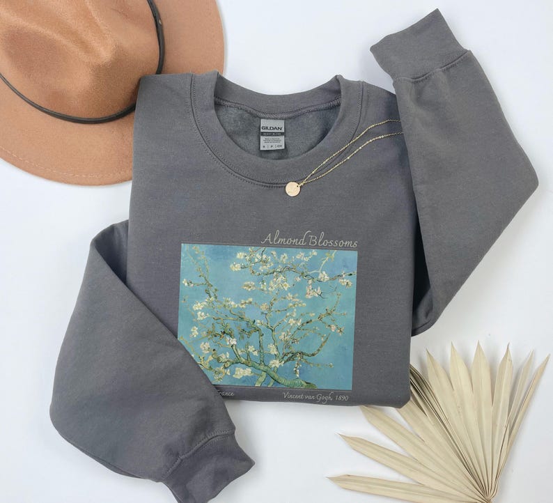 a gray sweatshirt with a graphic of a tree with white flowers, a brown hat, a necklace, and a fan on a white background.