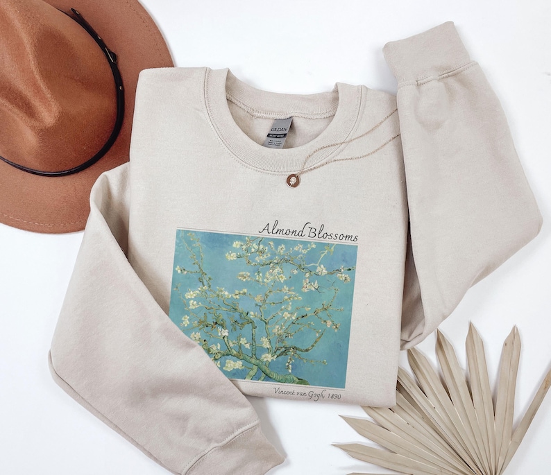 a beige sweatshirt with a printed design of a tree with white flowers, a brown hat, and a fan, all placed on a white background.