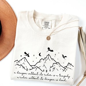 A white t-shirt with a graphic design depicting mountains and a dragon flying over them, along with a quote that reads "A dragon without its rider is a tragedy, a rider without its dragon is dead."